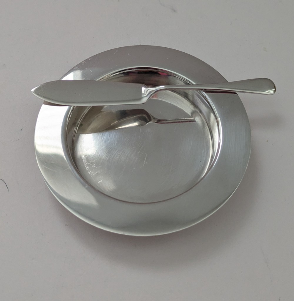 A boxed set of six circular sterling silver butter dishes paired with six matching butter knives inside a silk-lined presentation case. View X