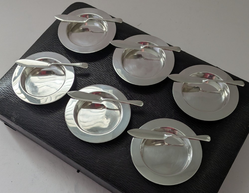 A boxed set of six circular sterling silver butter dishes paired with six matching butter knives inside a silk-lined presentation case. View XIV