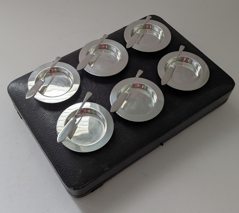 A boxed set of six circular sterling silver butter dishes paired with six matching butter knives inside a silk-lined presentation case. View XV