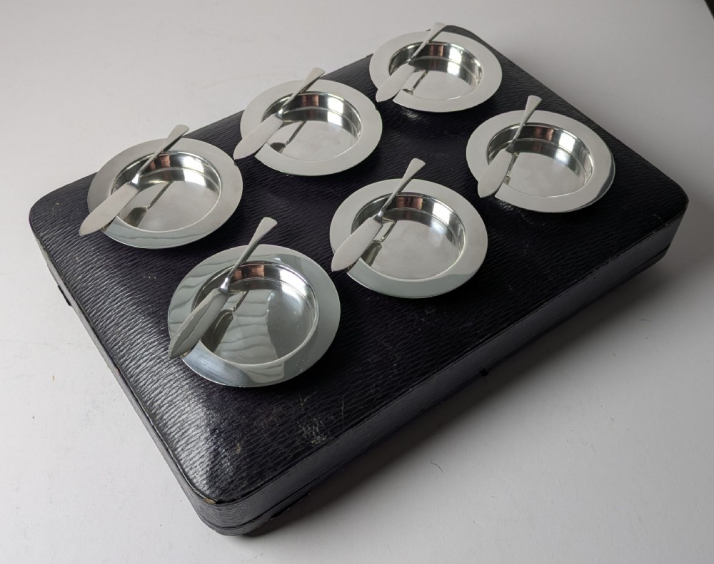 A boxed set of six circular sterling silver butter dishes paired with six matching butter knives inside a silk-lined presentation case. View XVI