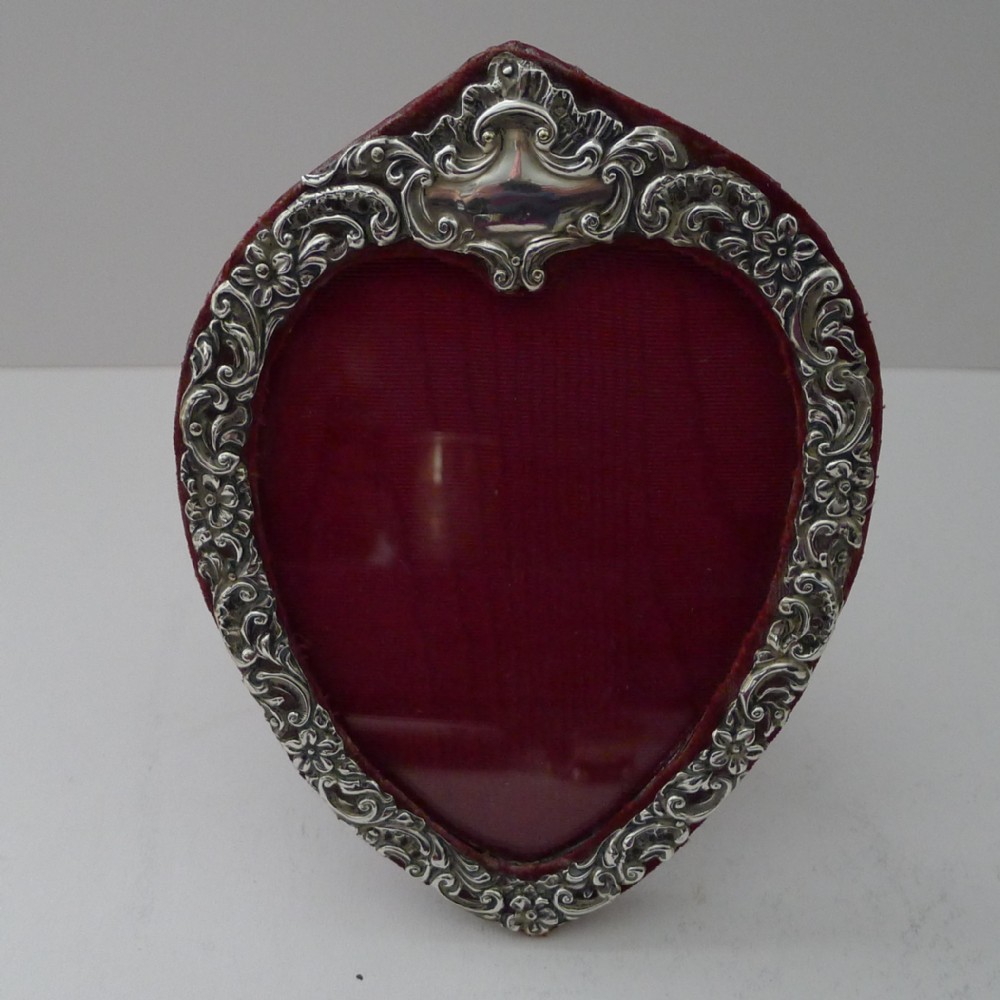 An Edwardian heart-shaped sterling silver photo frame with ornate scroll and floral repousse work set against a crimson velvet backing.