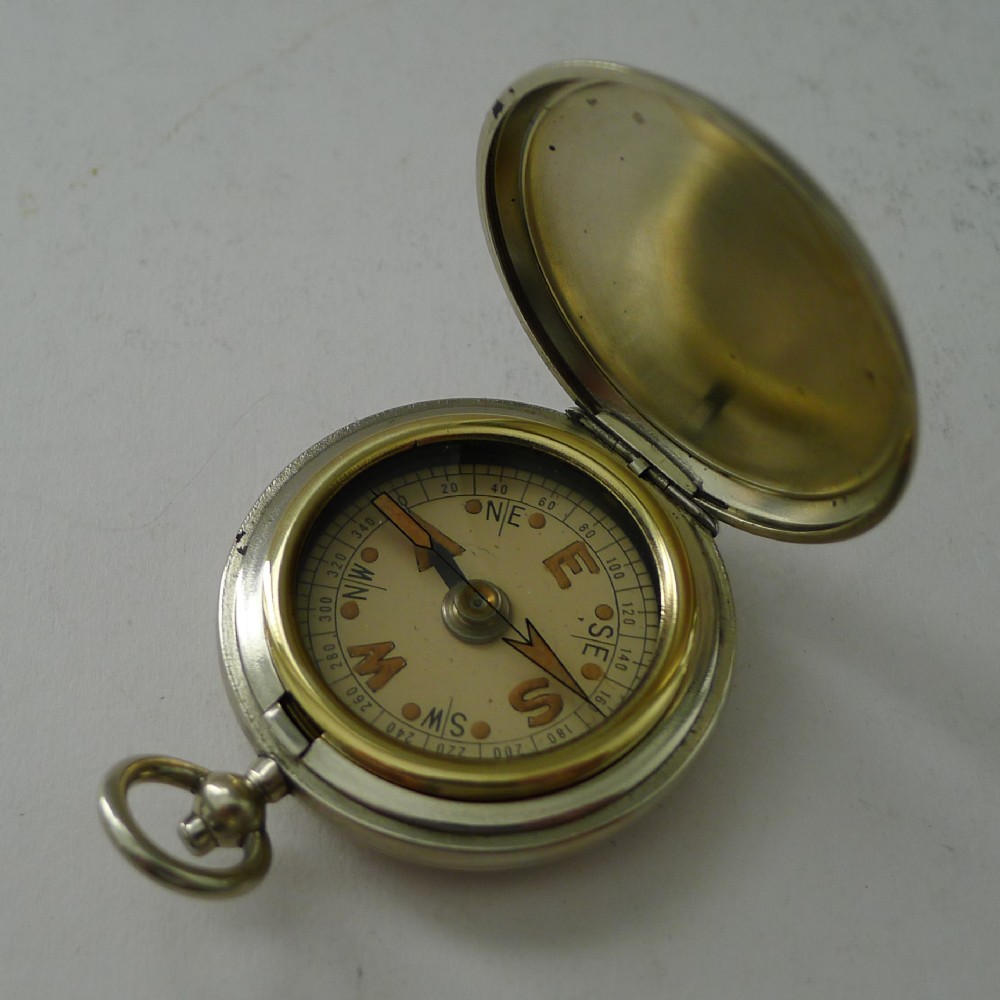 An opened antique pocket watch style military compass with a nickel casing and brass rim surrounding a cream dial.