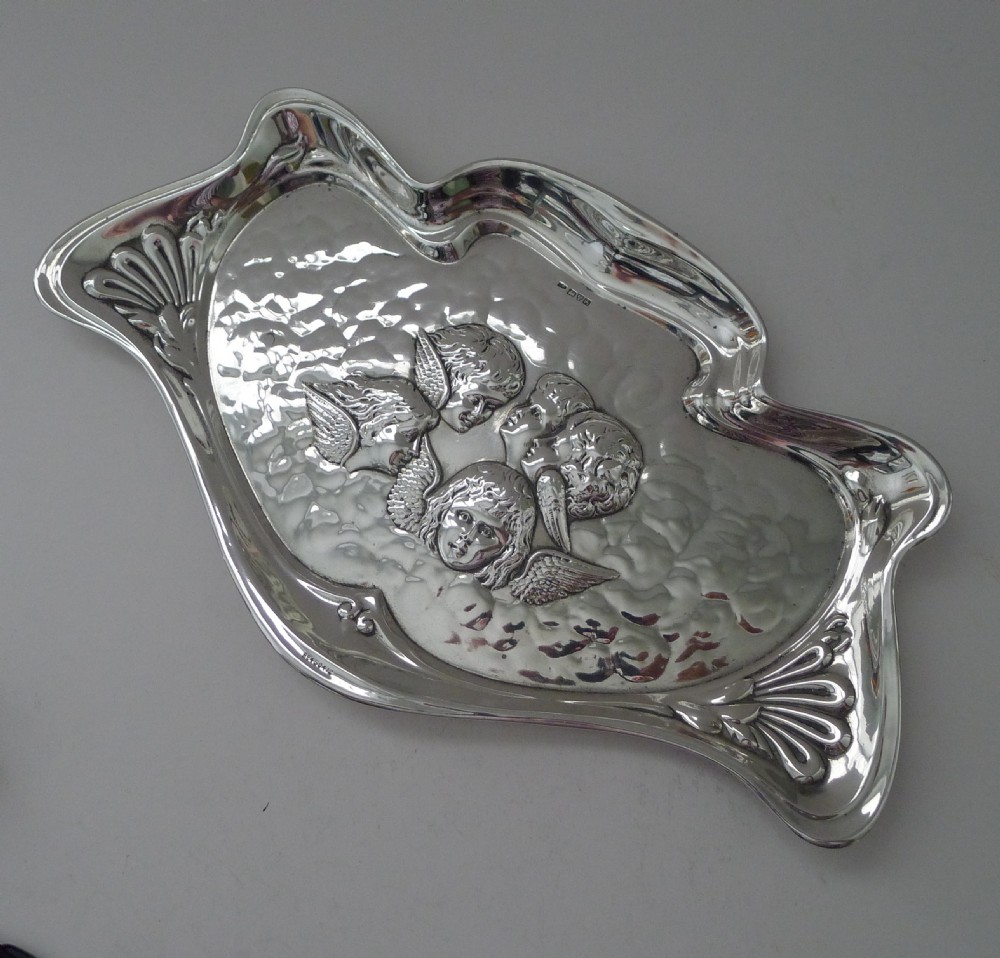 A shaped sterling silver dressing table tray featuring embossed cherub heads surrounded by clouds and stylized Art Nouveau scrolling borders. View V