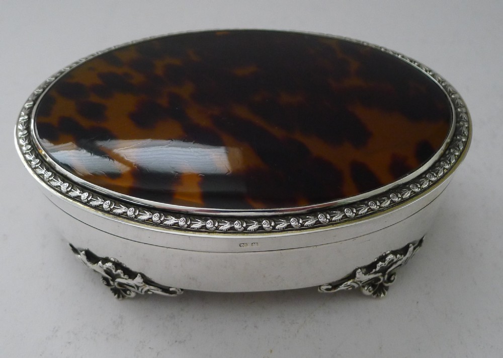 An oval sterling silver jewellery box featuring an inset tortoiseshell lid, decorative silver rim border, and four ornate cast feet. View VI