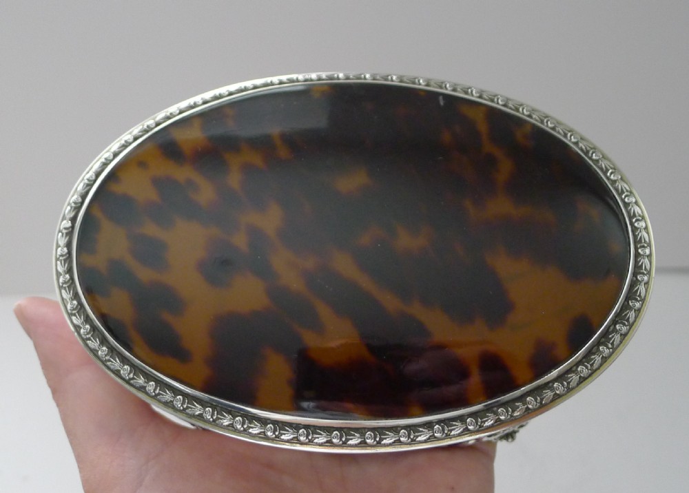 An oval sterling silver jewellery box featuring an inset tortoiseshell lid, decorative silver rim border, and four ornate cast feet. View XII