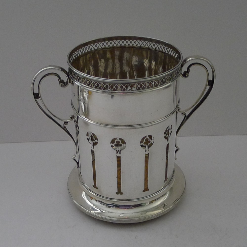 An Edwardian sterling silver wine bottle holder featuring pierced vertical motifs, a lattice rim, dual scroll handles, and a circular base.