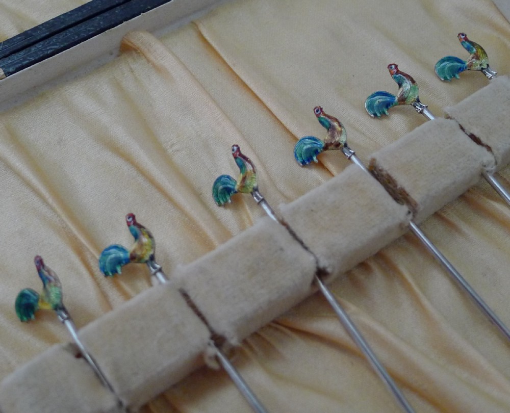 A set of six sterling silver cocktail sticks featuring colourful enamelled cockerel finials inside a silk-lined presentation box.