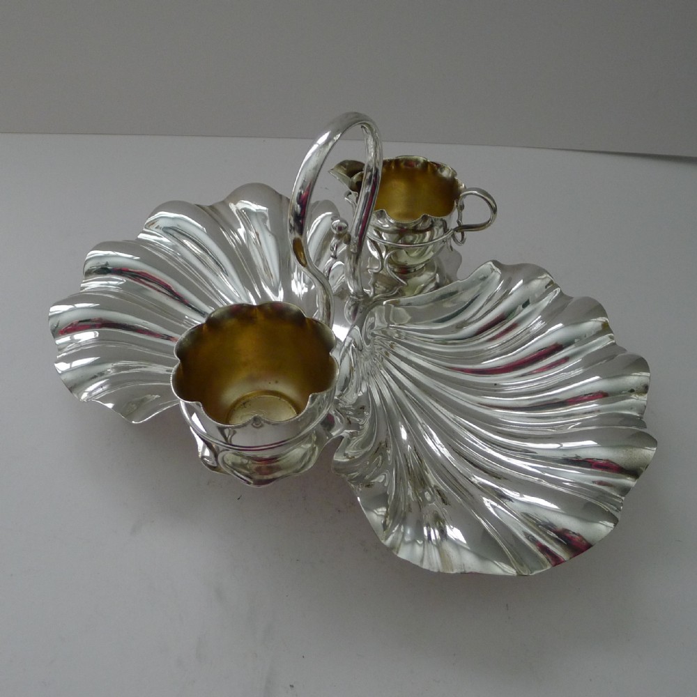 An antique silver-plated strawberry set featuring two fluted shell-shaped serving dishes, a central overhead loop handle, and two small gilt-lined pots.