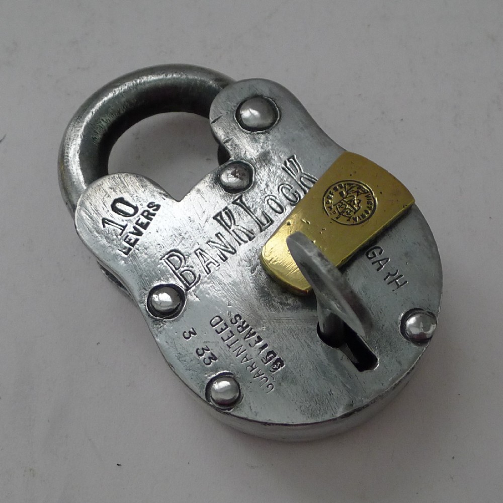 Heavy steel bank padlock featuring stamped lettering and a pivoting brass keyhole cover with an inserted key.