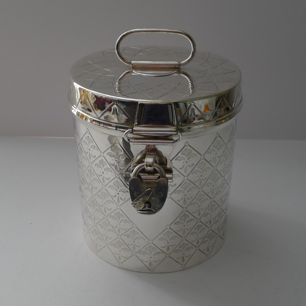 A cylindrical silver-plated Victorian biscuit box featuring engraved geometric trellis patterning, a loop top handle, and a hinged clasp with a padlock.