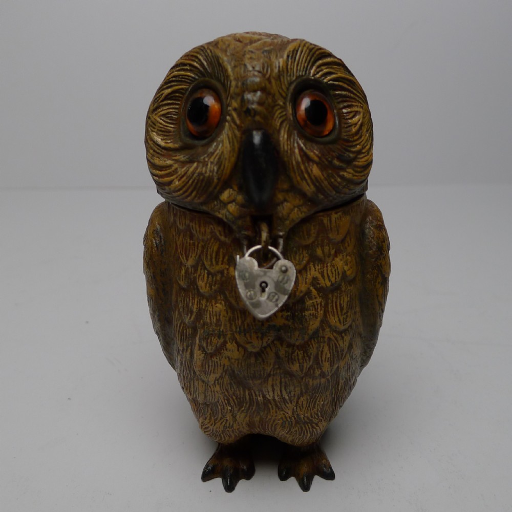 A painted spelter money box shaped as an owl featuring amber glass eyes and a small heart-shaped lock motif at its throat.