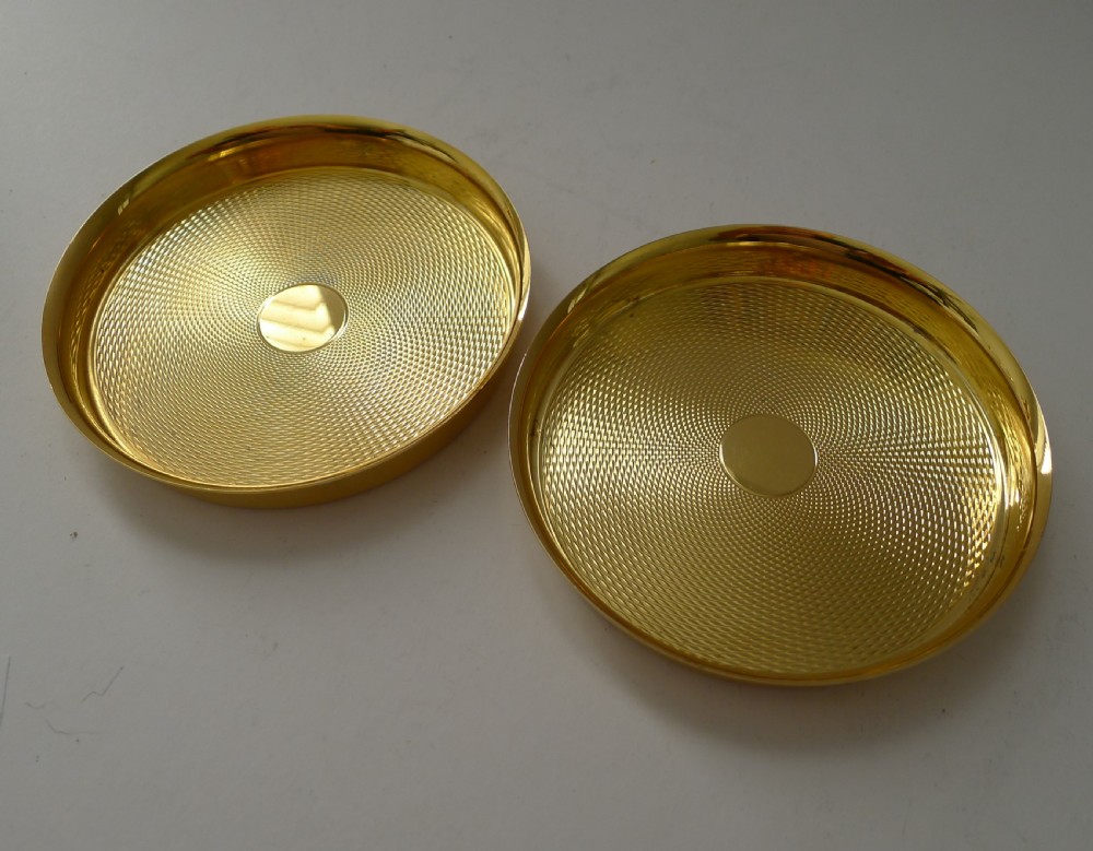 A pair of round gold-coloured vermeil silver gilt coasters featuring engine-turned textured centres and smooth raised rims. View VI