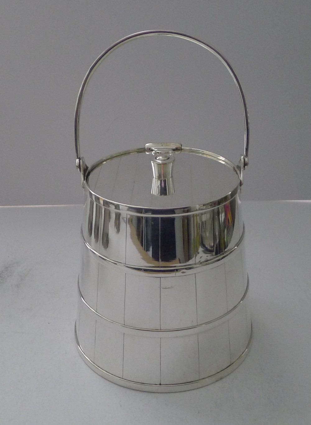 A silver-plated cylindrical container styled as a coopered wooden bucket with incised vertical lines, horizontal bands, a flat lid, and a tall curved swing handle.