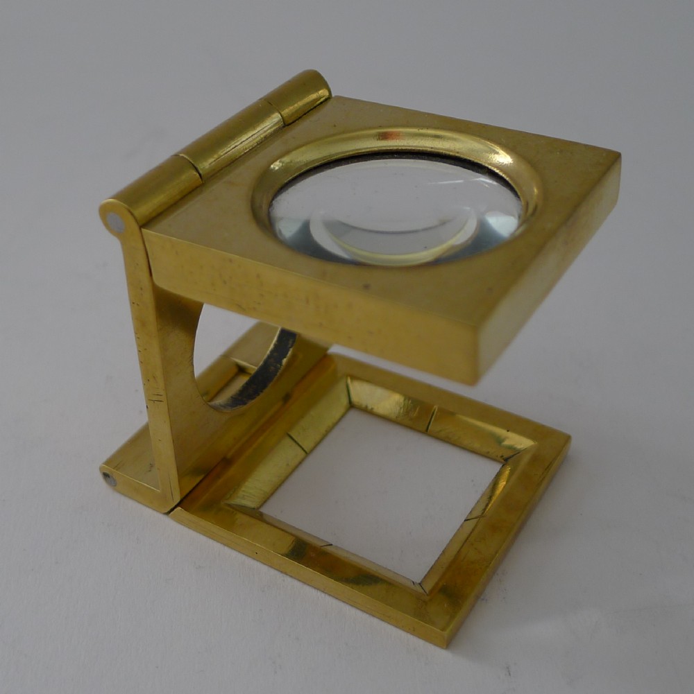 An antique brass folding linen thread counter with a circular magnifying lens held in a hinged frame above a square measurement aperture.