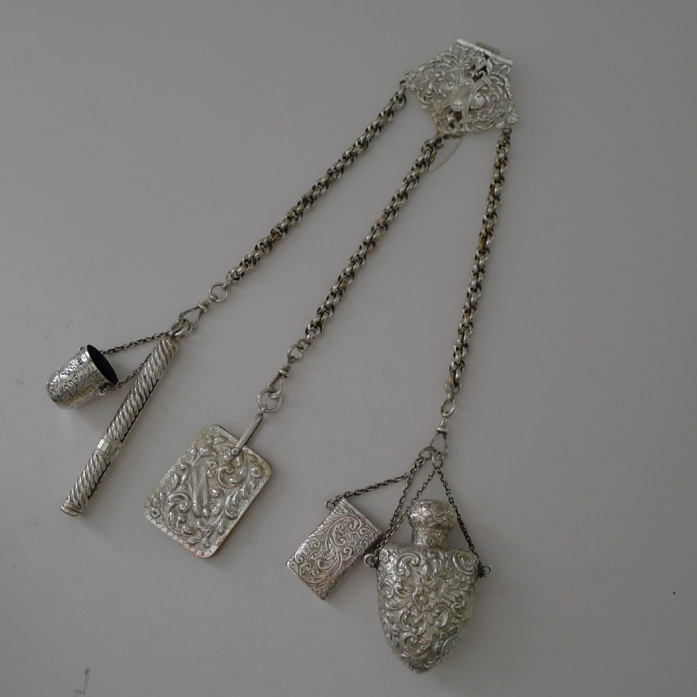 A Victorian sterling silver chatelaine featuring an ornate pierced scroll waist clip suspending five linked chains attached to silver accessories including a scent bottle, card case, pencil, and thimble holder.