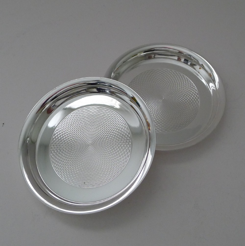 A pair of circular silver-plated coasters featuring raised rims and engine-turned concentric circle patterns.