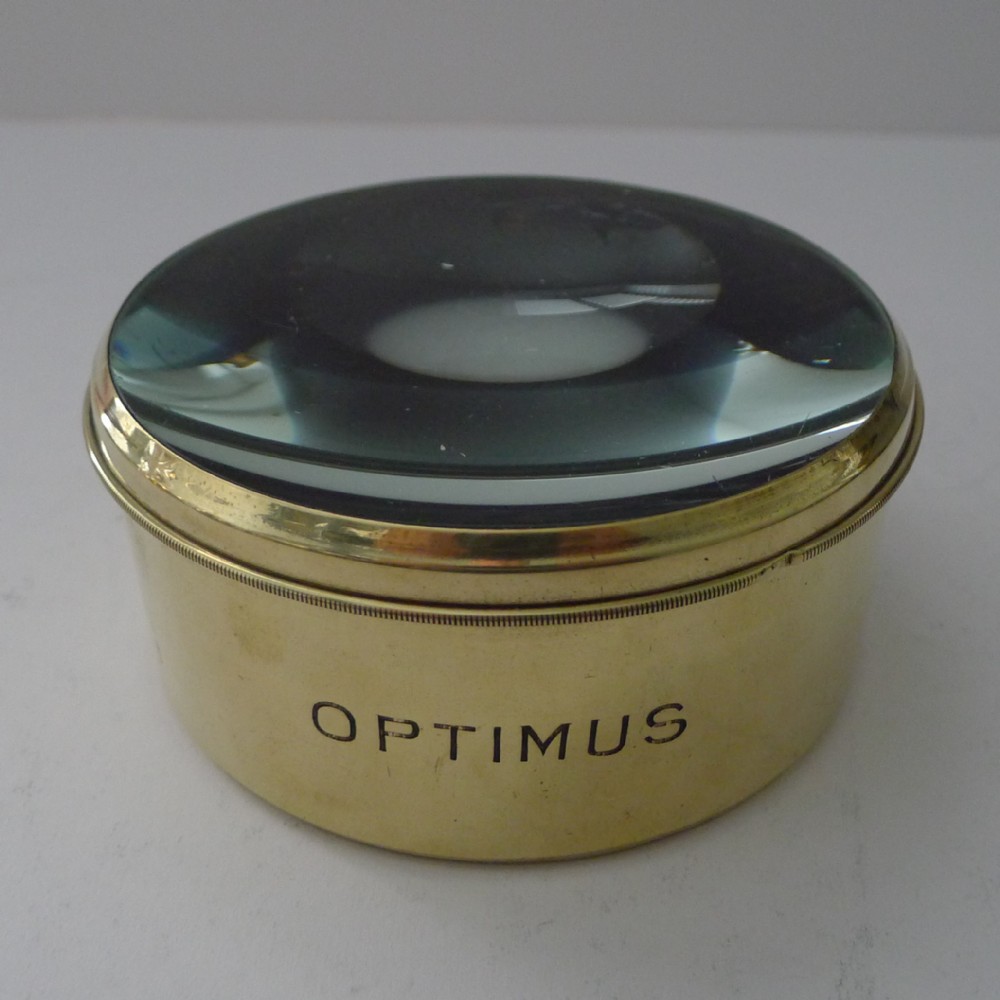 A cylindrical brass-bound magnifying glass paperweight engraved with the name Optimus around its base.