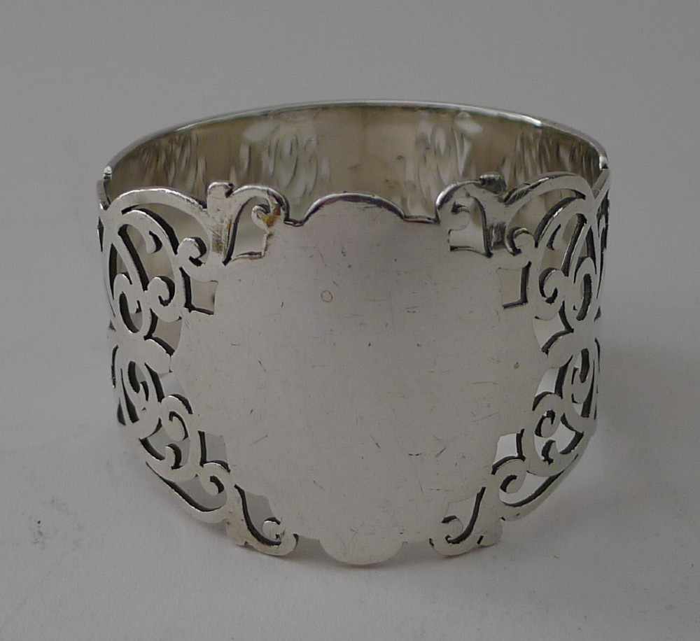 A cylindrical sterling silver napkin ring featuring openwork scroll patterns and an unengraved shield cartouche.