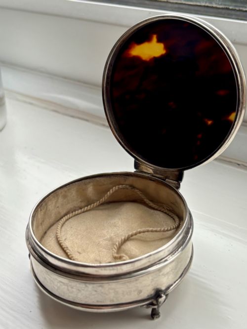 fig 1. A circular silver dressing table box featuring a dark tortoiseshell lid with silver floral basket inlay resting on small shaped feet.
