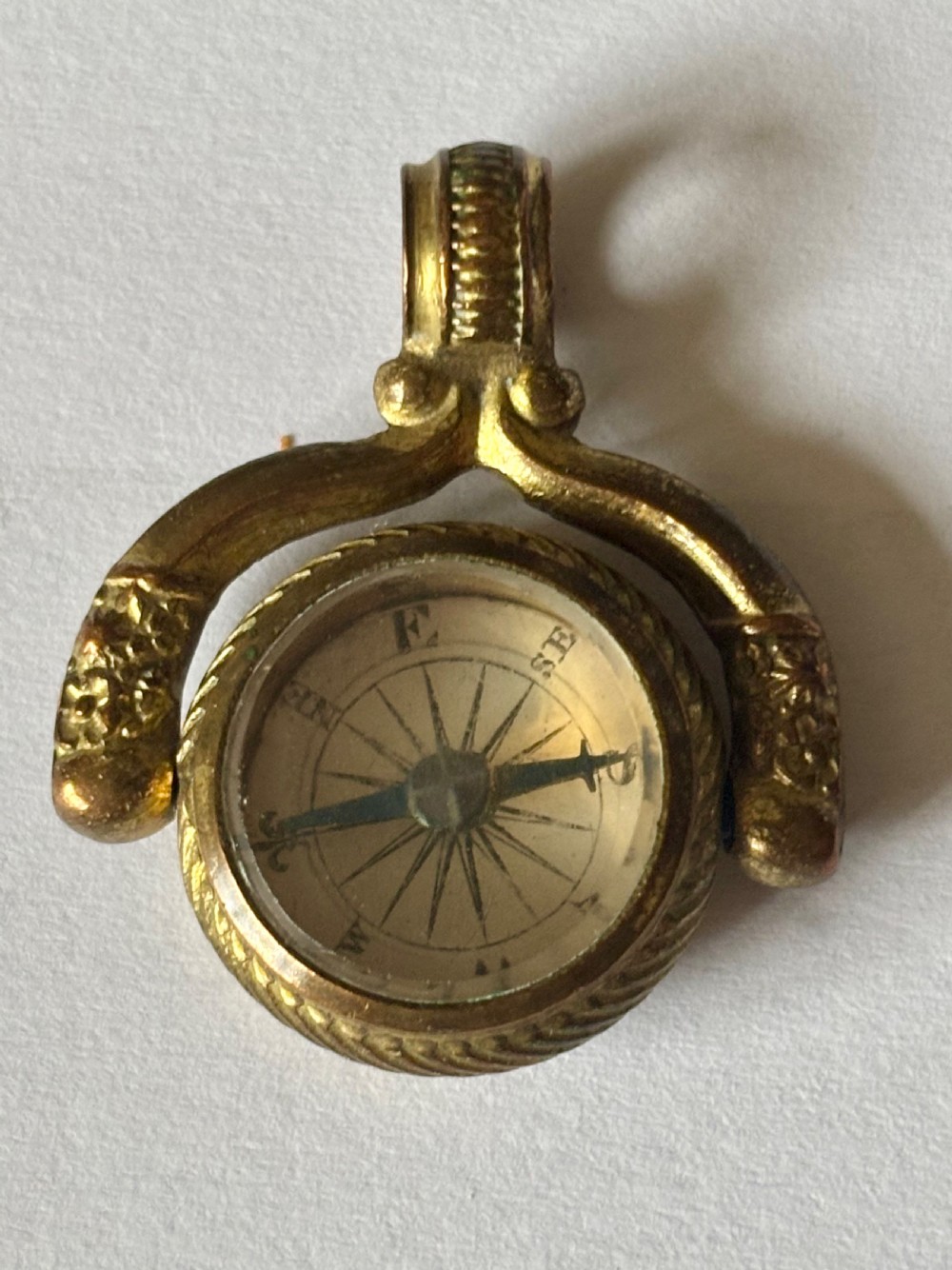An antique Victorian gilt metal swivel compass fob pendant featuring a textured suspension arch, a rope-twist border, and a compass dial beneath glass.