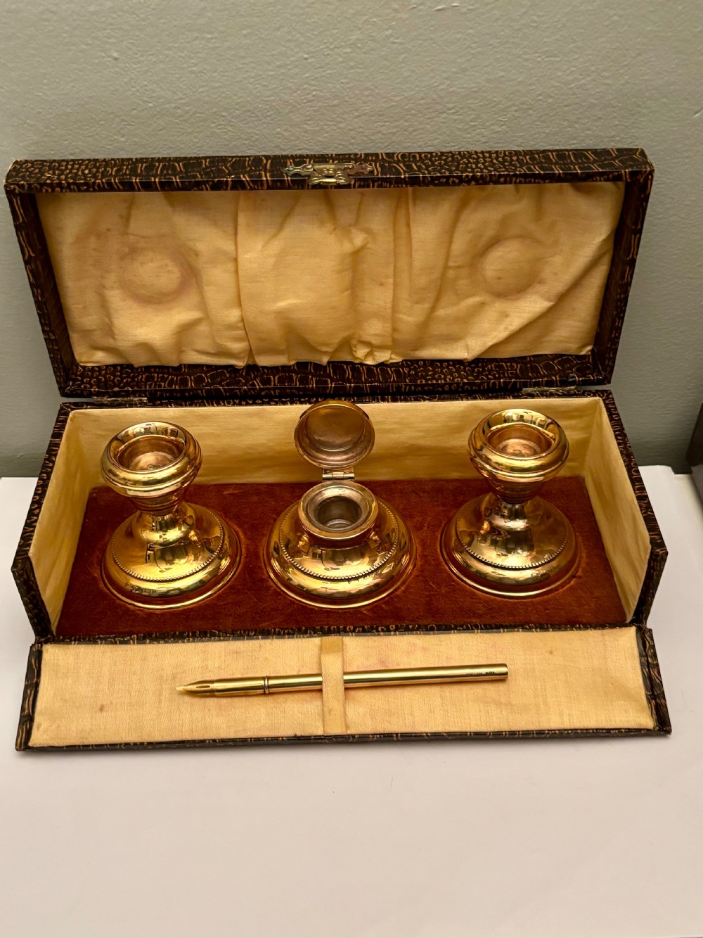 An open fitted presentation box containing two silver gilt candlesticks, a central inkwell, and a dip pen.