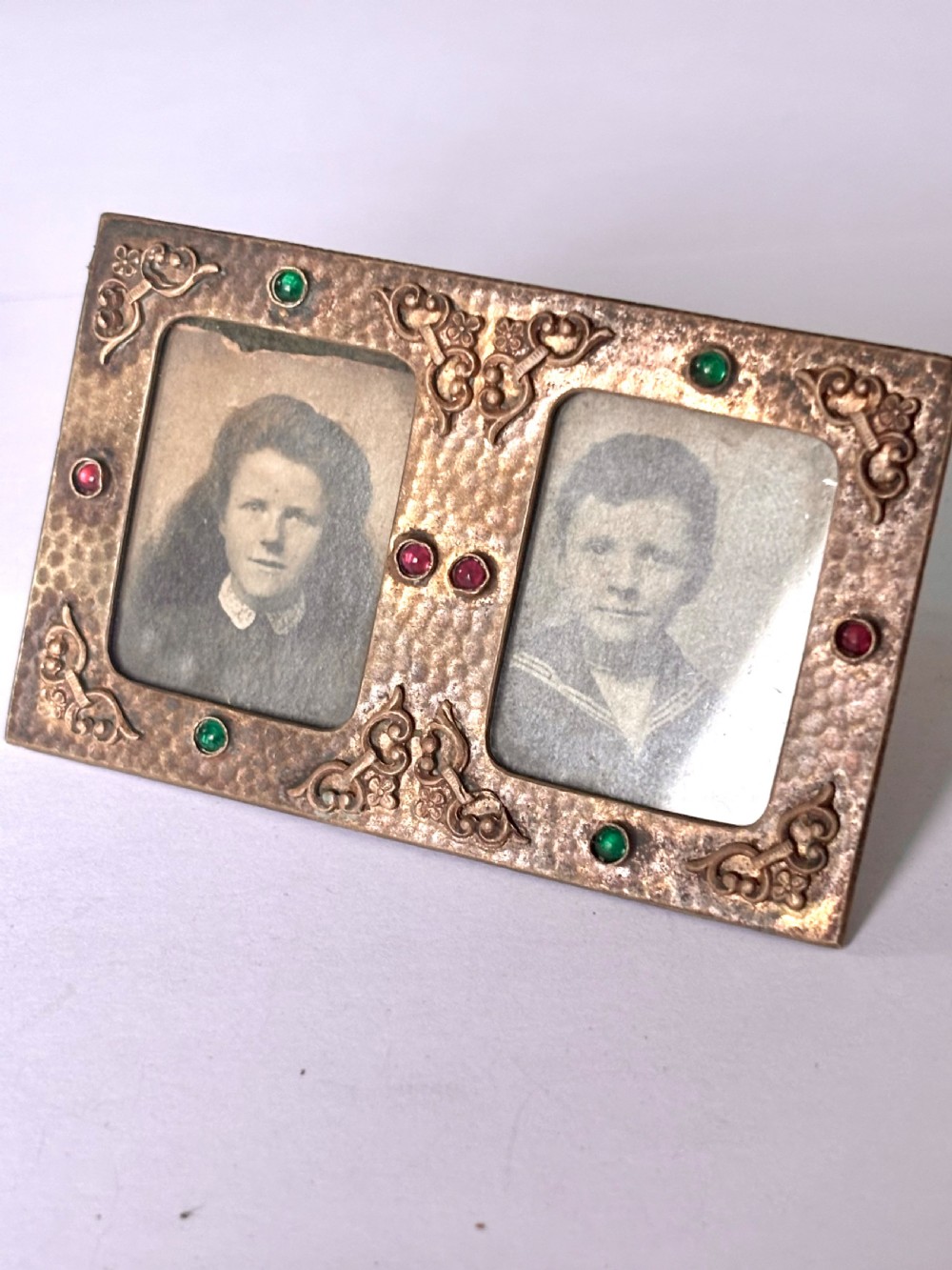 An antique Arts and Crafts double photo frame crafted from hammered gilt brass with applied scroll ornament and colourful cabochon stones containing two black and white portraits.