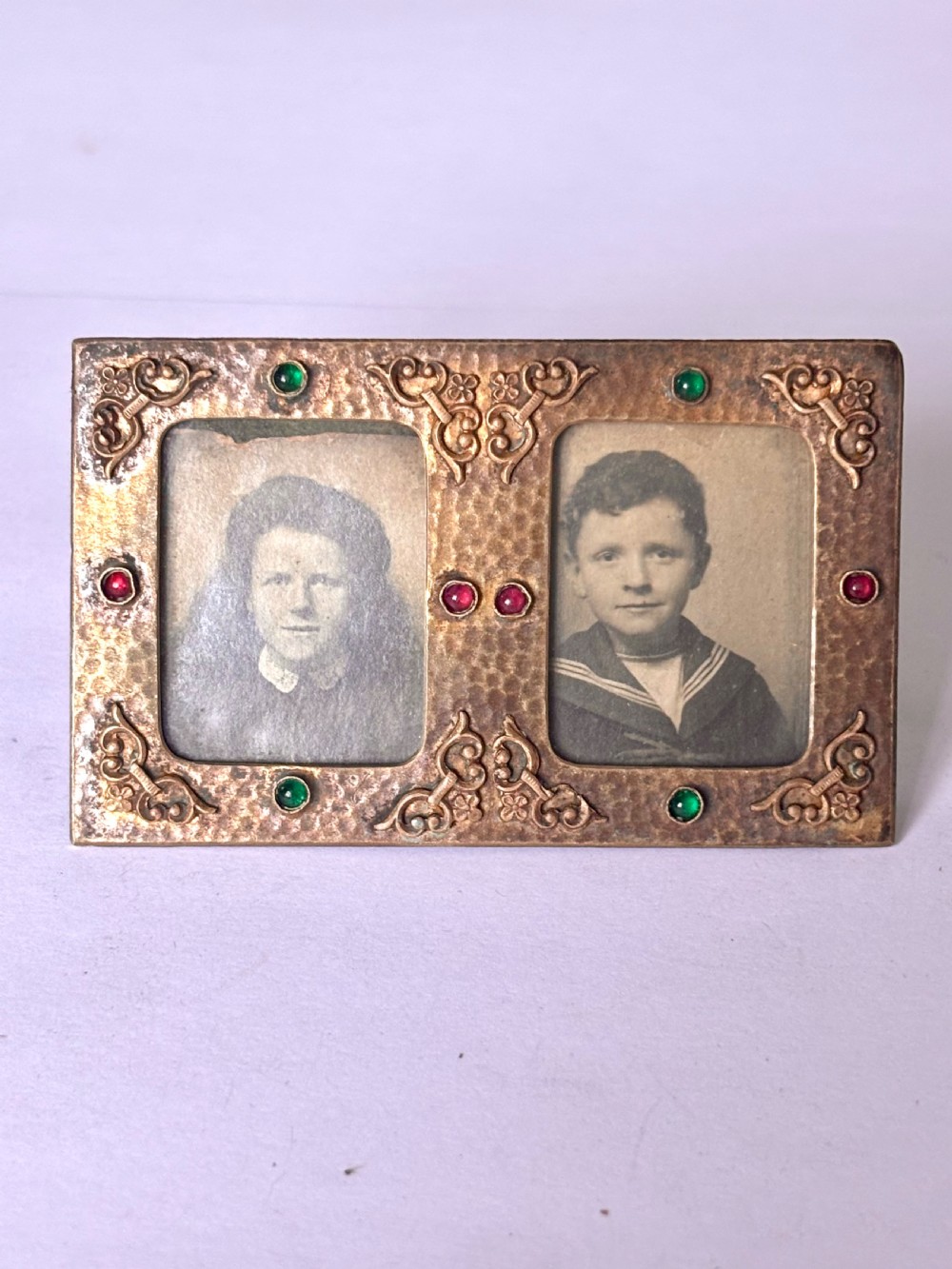 An antique Arts and Crafts double photo frame crafted from hammered gilt brass with applied scroll ornament and colourful cabochon stones containing two black and white portraits. View II