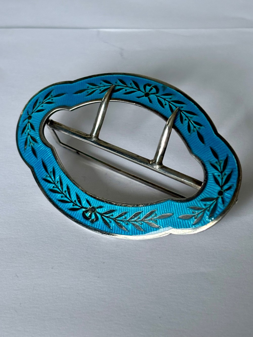 An oval Edwardian sterling silver belt buckle decorated with turquoise blue guilloche enamel and black leaf motifs.