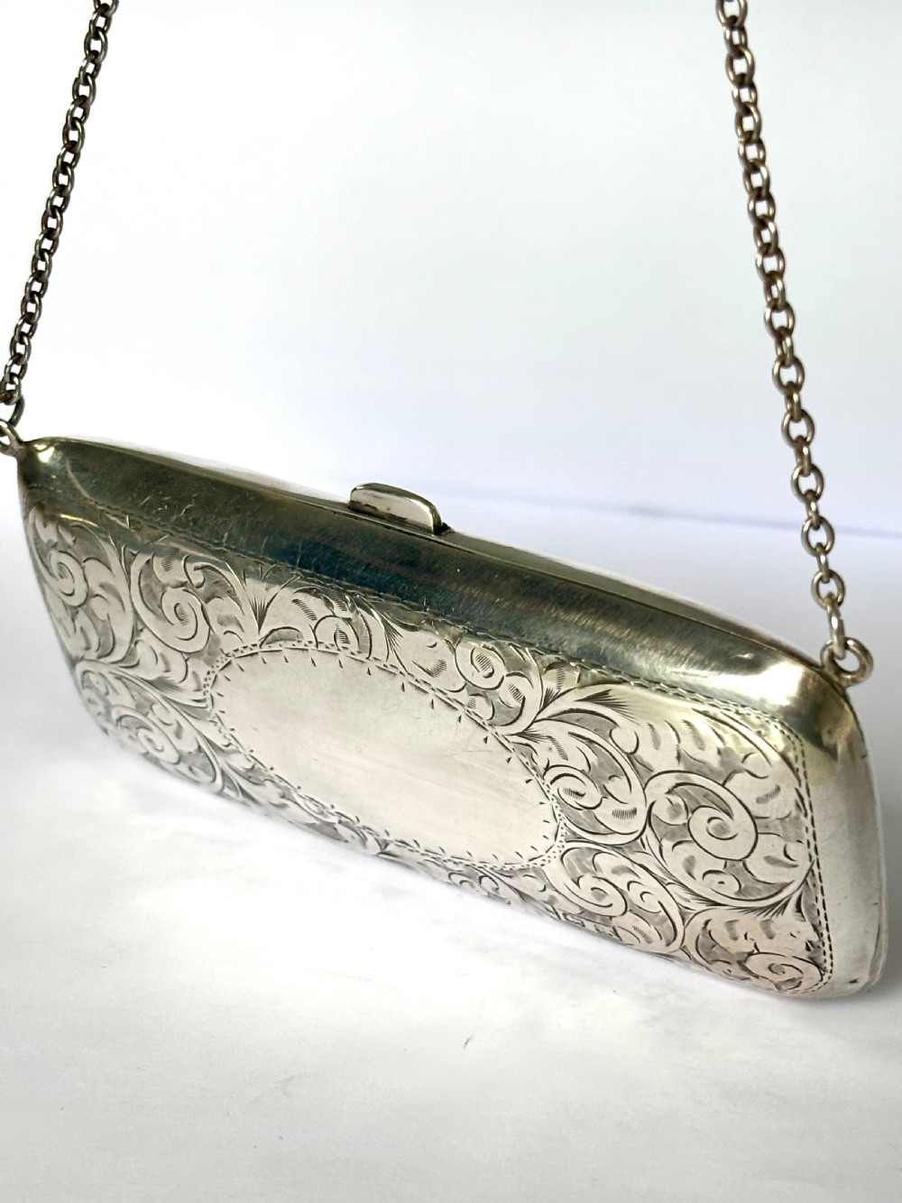An engraved Art Deco sterling silver purse featuring a fine chain handle, detailed floral scrollwork, and a central polished oval cartouche.