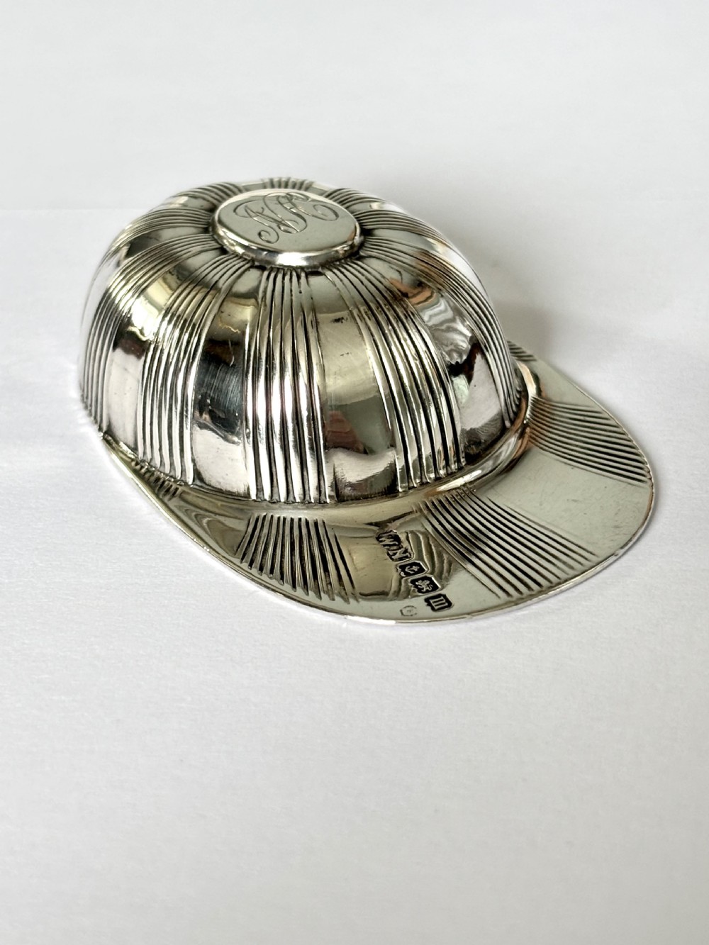 A solid sterling silver caddy spoon shaped like a peaked jockey cap with fluted panel detailing and British hallmarks along the brim.