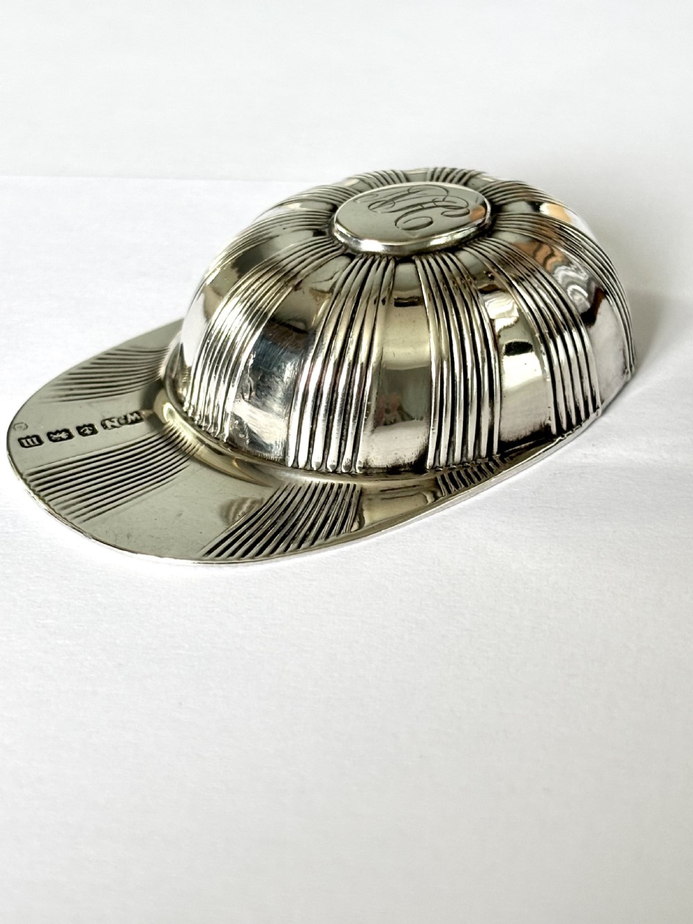 A solid sterling silver caddy spoon shaped like a peaked jockey cap with fluted panel detailing and British hallmarks along the brim. View II