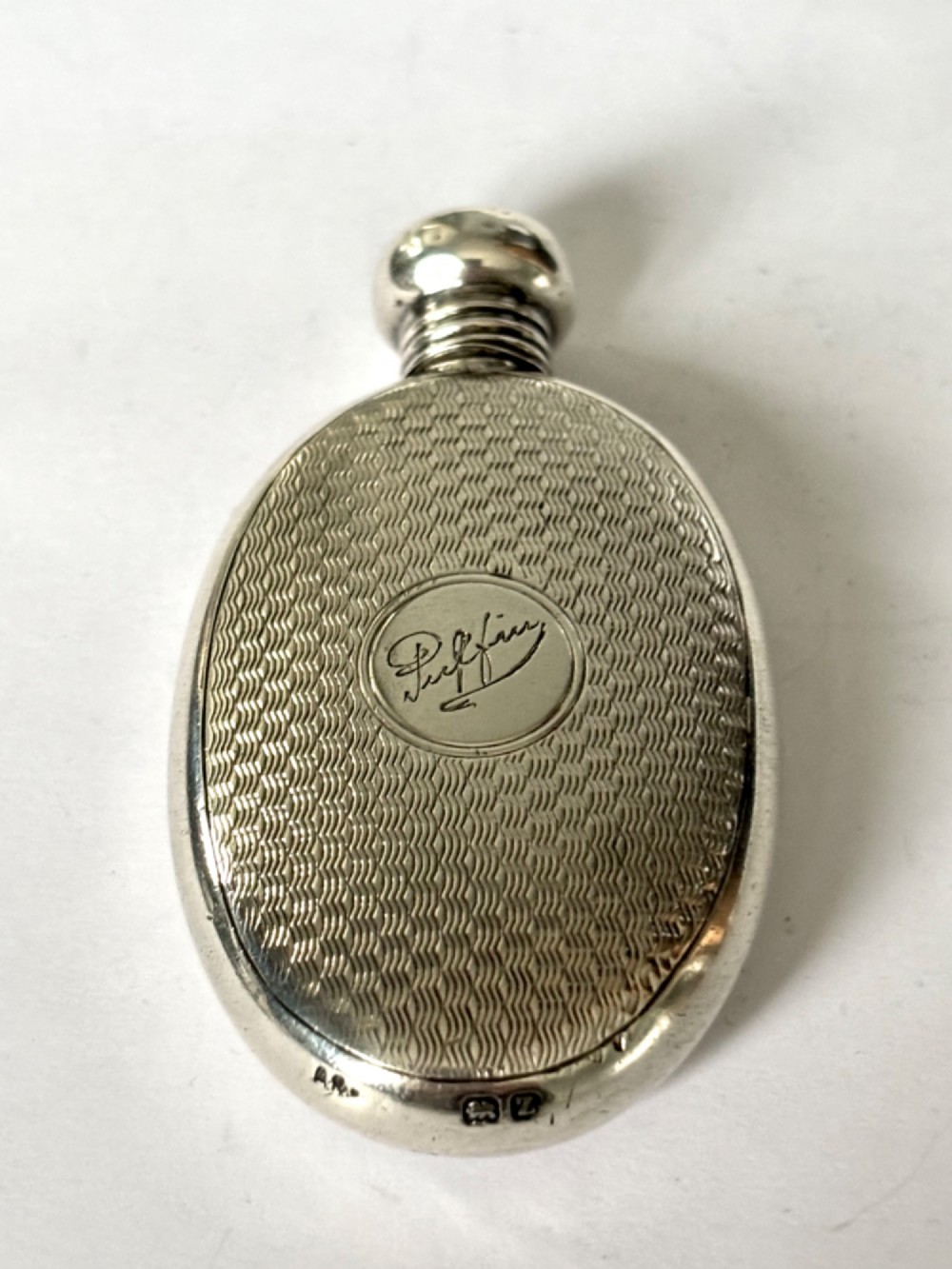 An oval sterling silver scent flask featuring an engine-turned pattern, a central engraved cartouche, a screw top cap, and British hallmarks at the base.