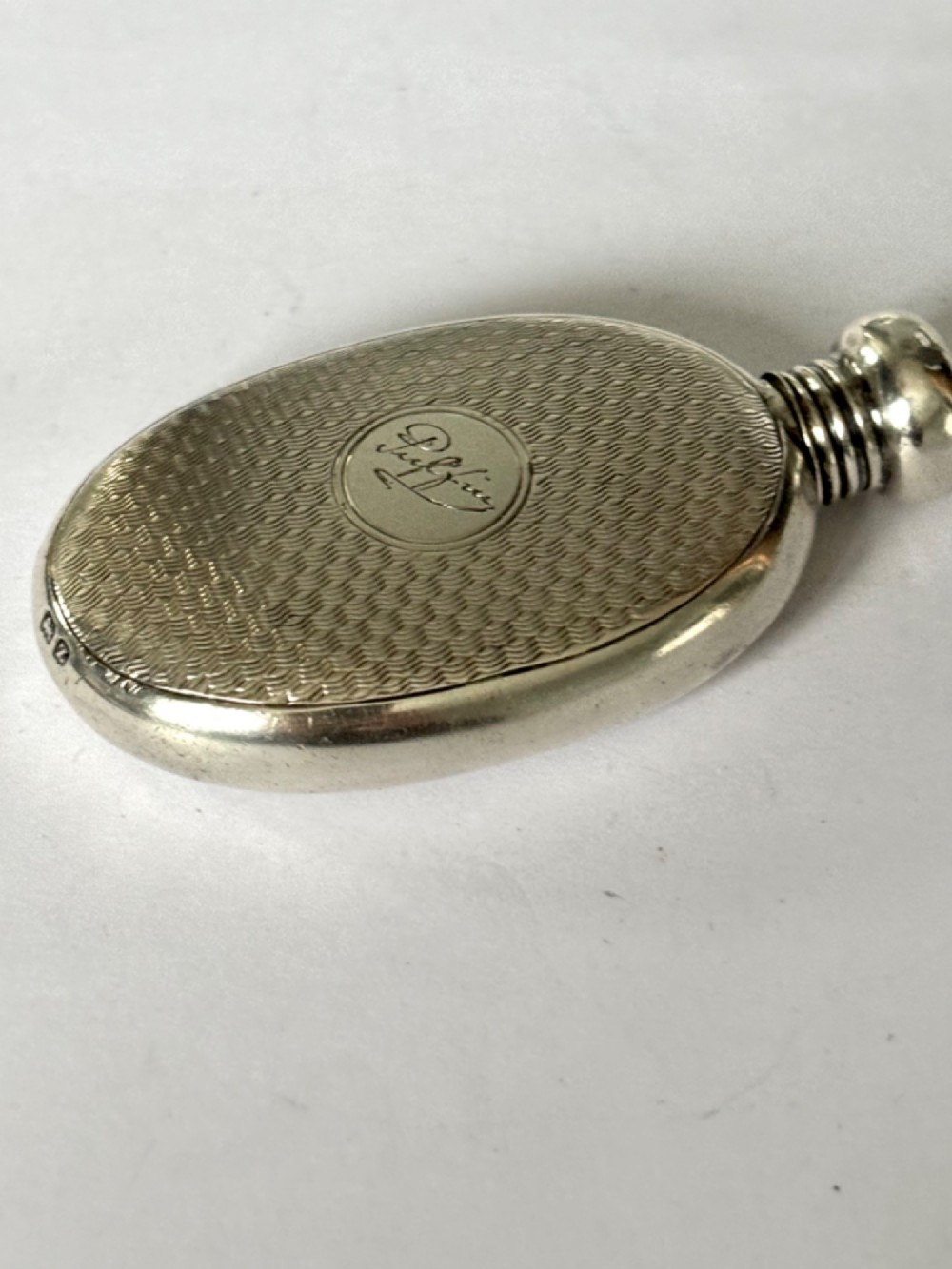 An oval sterling silver scent flask featuring an engine-turned pattern, a central engraved cartouche, a screw top cap, and British hallmarks at the base. View II