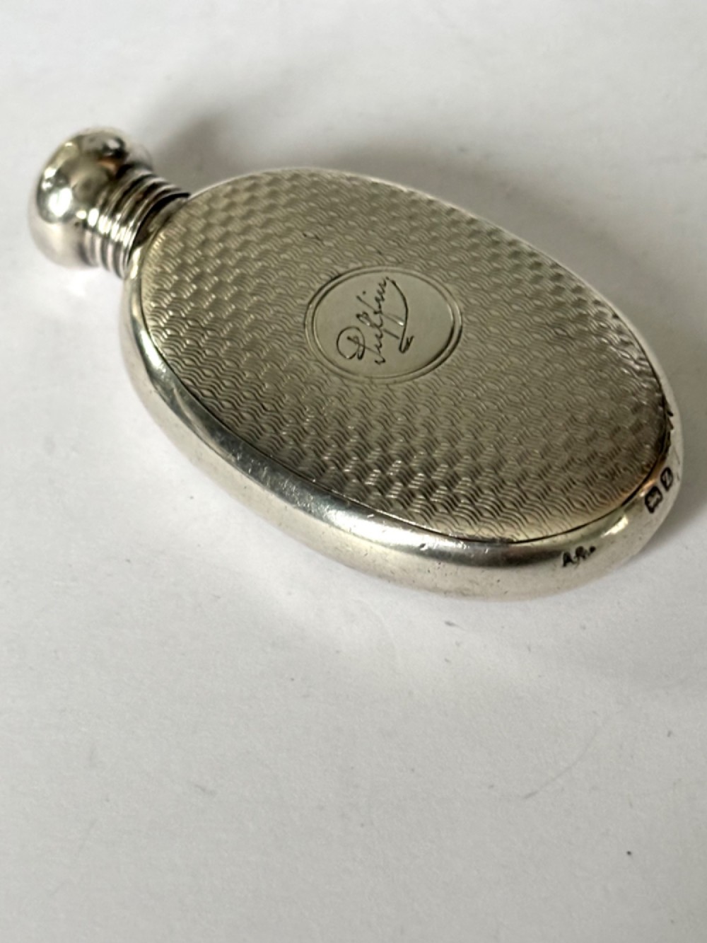 An oval sterling silver scent flask featuring an engine-turned pattern, a central engraved cartouche, a screw top cap, and British hallmarks at the base. View III