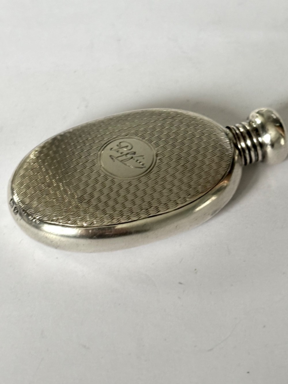 An oval sterling silver scent flask featuring an engine-turned pattern, a central engraved cartouche, a screw top cap, and British hallmarks at the base. View IV
