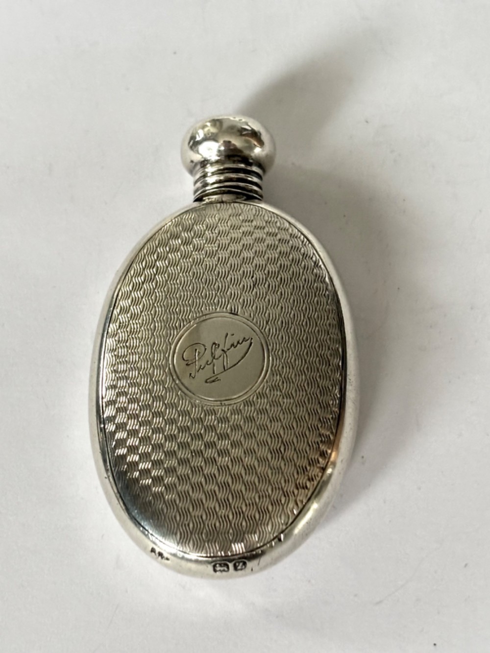 An oval sterling silver scent flask featuring an engine-turned pattern, a central engraved cartouche, a screw top cap, and British hallmarks at the base. View V