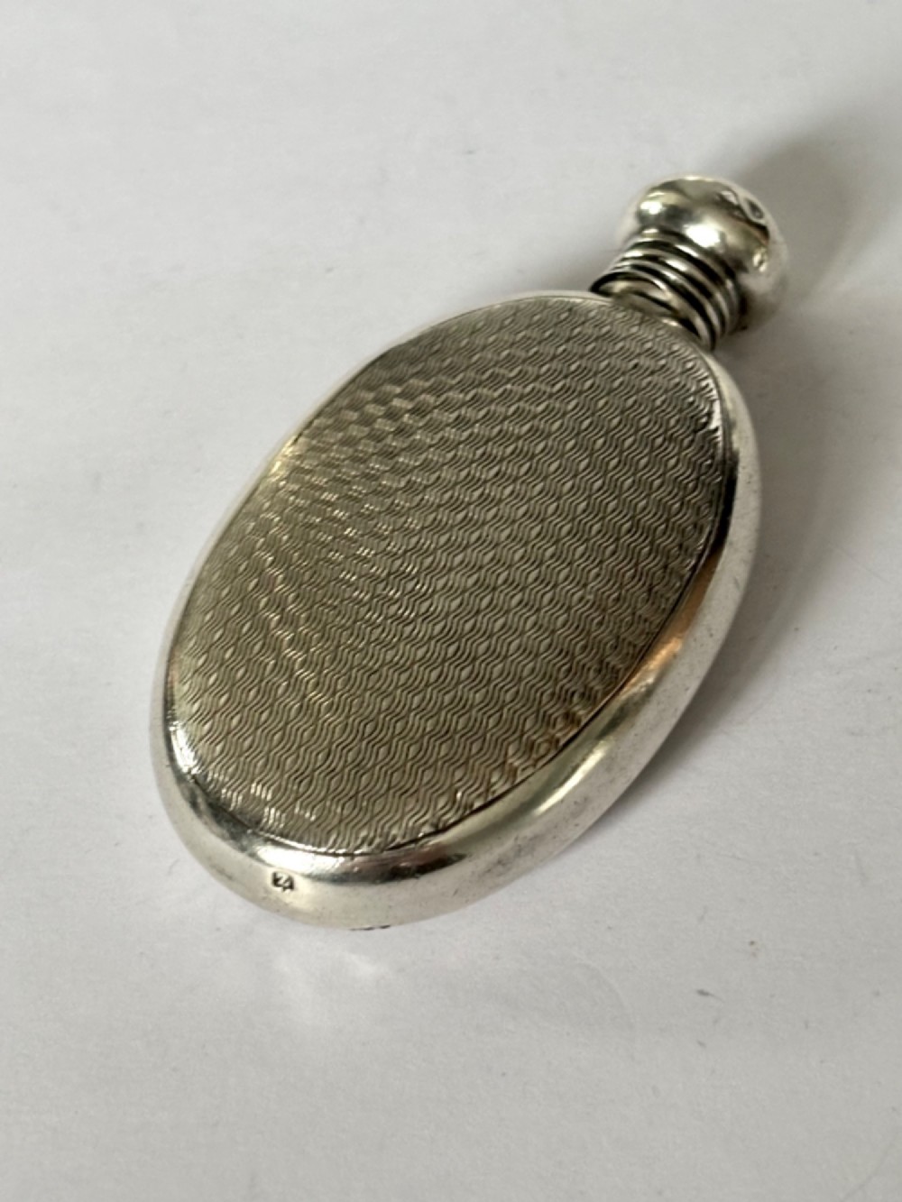 An oval sterling silver scent flask featuring an engine-turned pattern, a central engraved cartouche, a screw top cap, and British hallmarks at the base. View VI