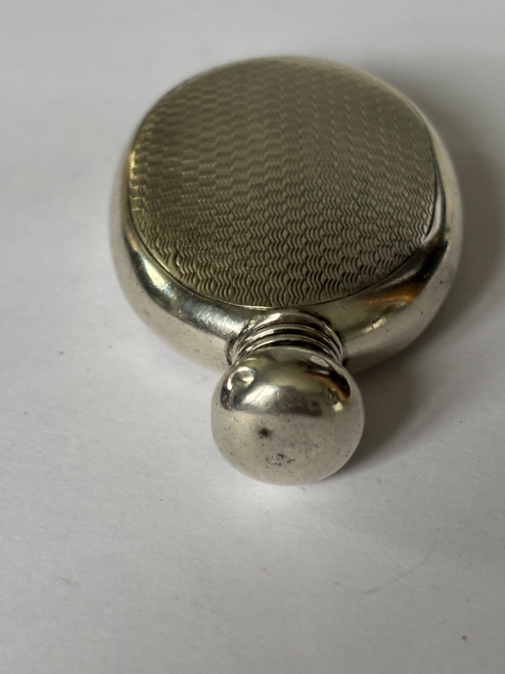 An oval sterling silver scent flask featuring an engine-turned pattern, a central engraved cartouche, a screw top cap, and British hallmarks at the base. View VII