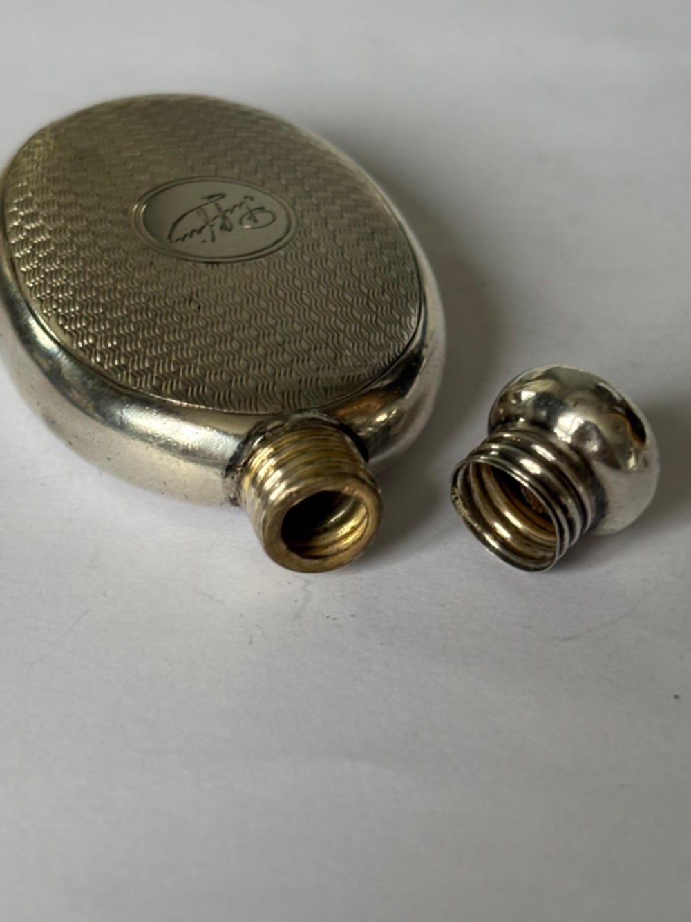 An oval sterling silver scent flask featuring an engine-turned pattern, a central engraved cartouche, a screw top cap, and British hallmarks at the base. View IX