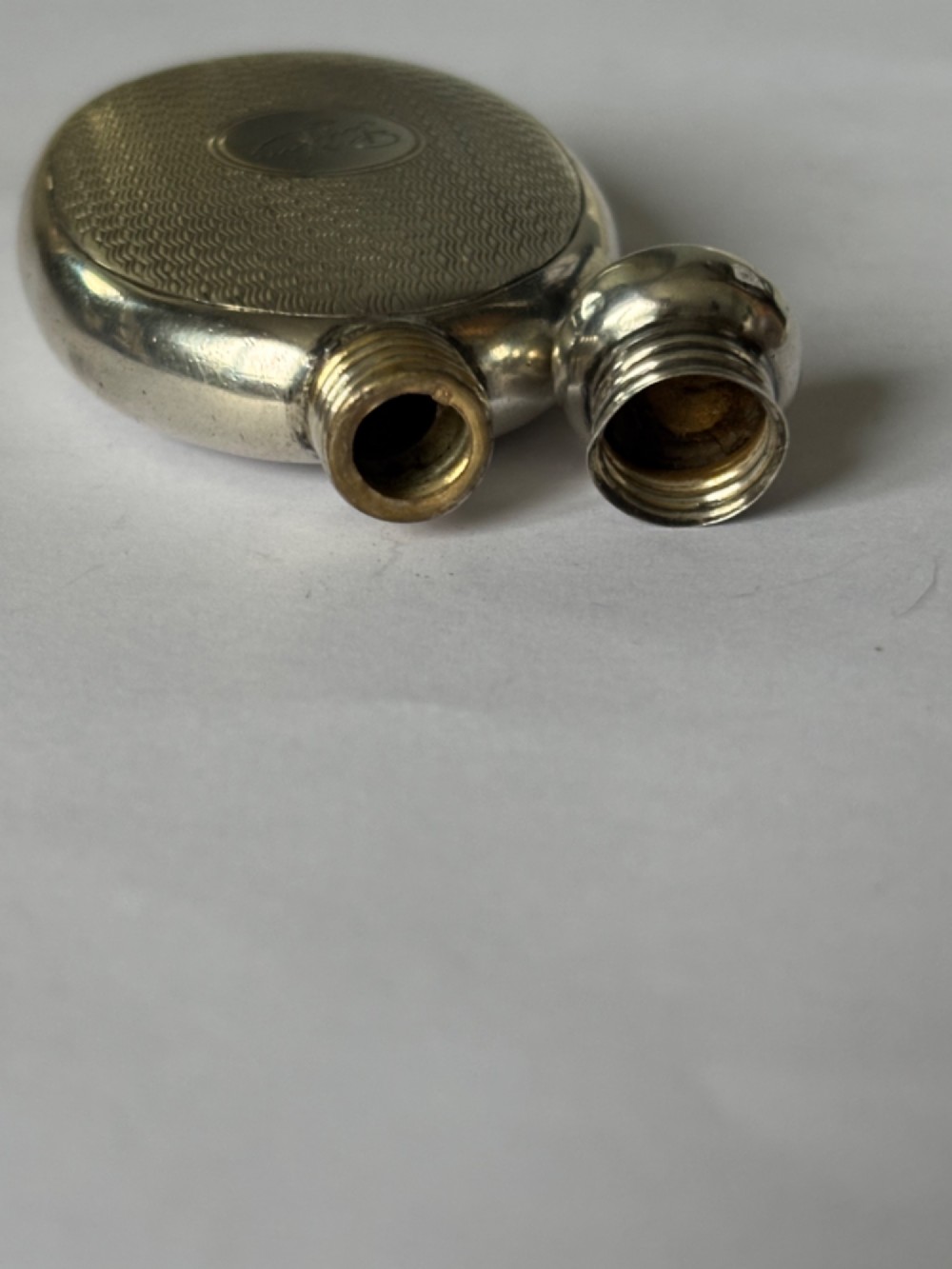 An oval sterling silver scent flask featuring an engine-turned pattern, a central engraved cartouche, a screw top cap, and British hallmarks at the base. View X