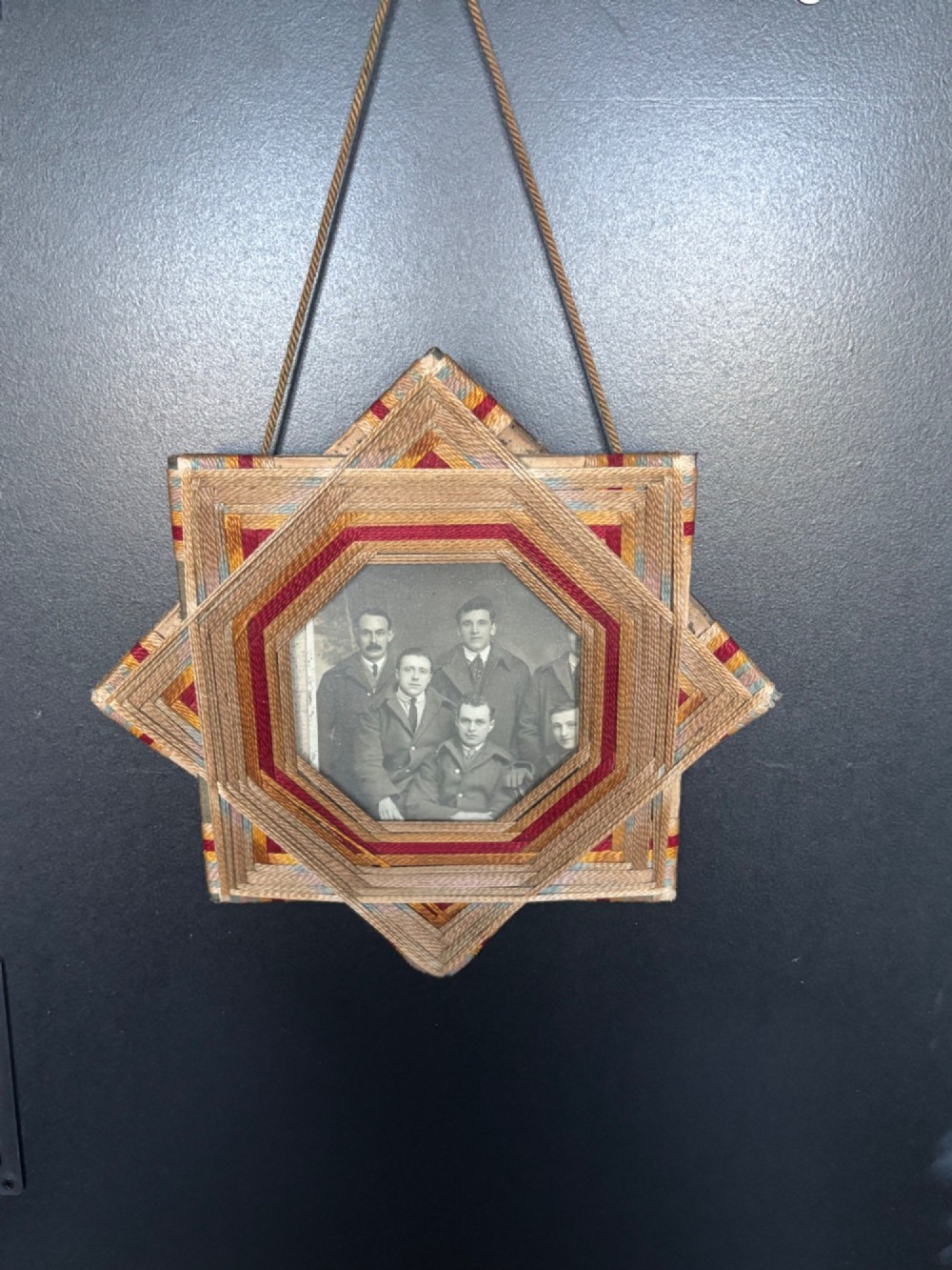 An octagram star-shaped folk art picture frame wrapped in beige, red, and gold string, surrounding a monochrome portrait photograph of five men.