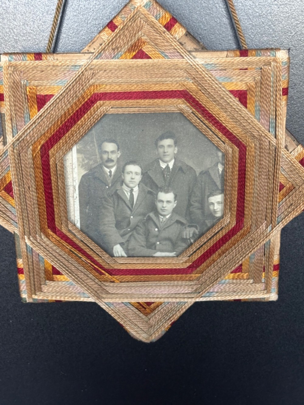 An octagram star-shaped folk art picture frame wrapped in beige, red, and gold string, surrounding a monochrome portrait photograph of five men. View III