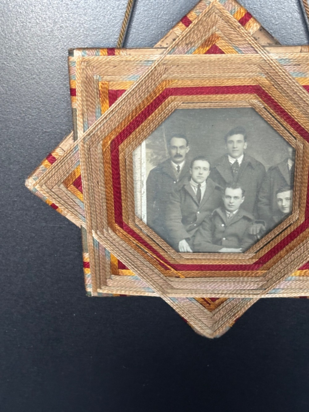 An octagram star-shaped folk art picture frame wrapped in beige, red, and gold string, surrounding a monochrome portrait photograph of five men. View IV