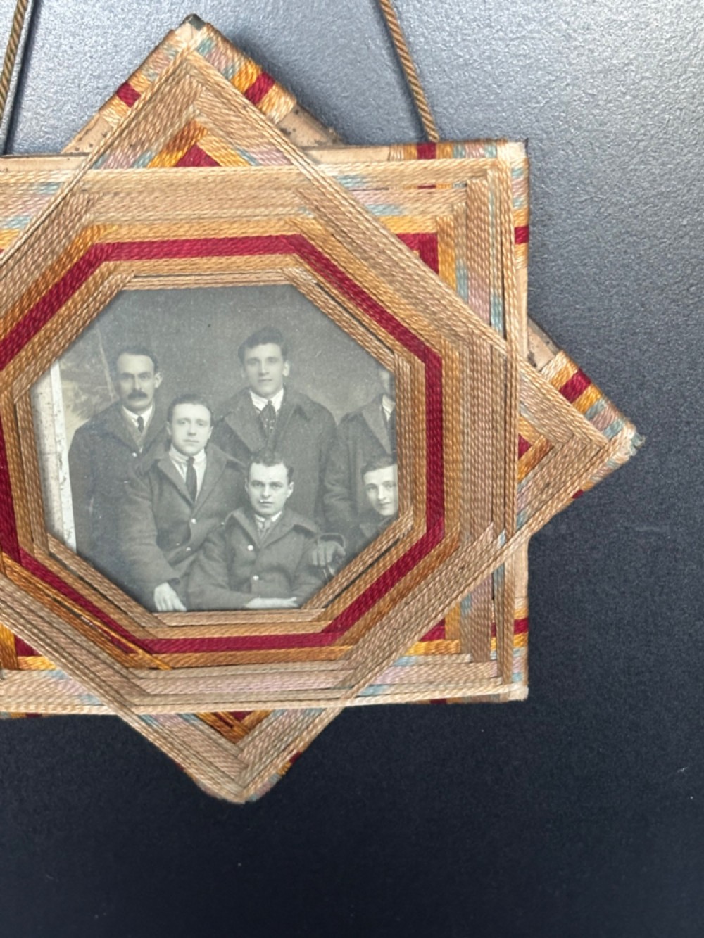 An octagram star-shaped folk art picture frame wrapped in beige, red, and gold string, surrounding a monochrome portrait photograph of five men. View V