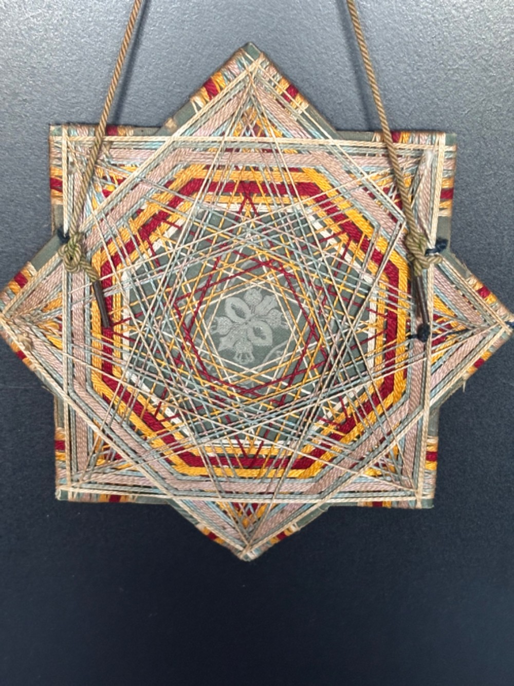 An octagram star-shaped folk art picture frame wrapped in beige, red, and gold string, surrounding a monochrome portrait photograph of five men. View VI