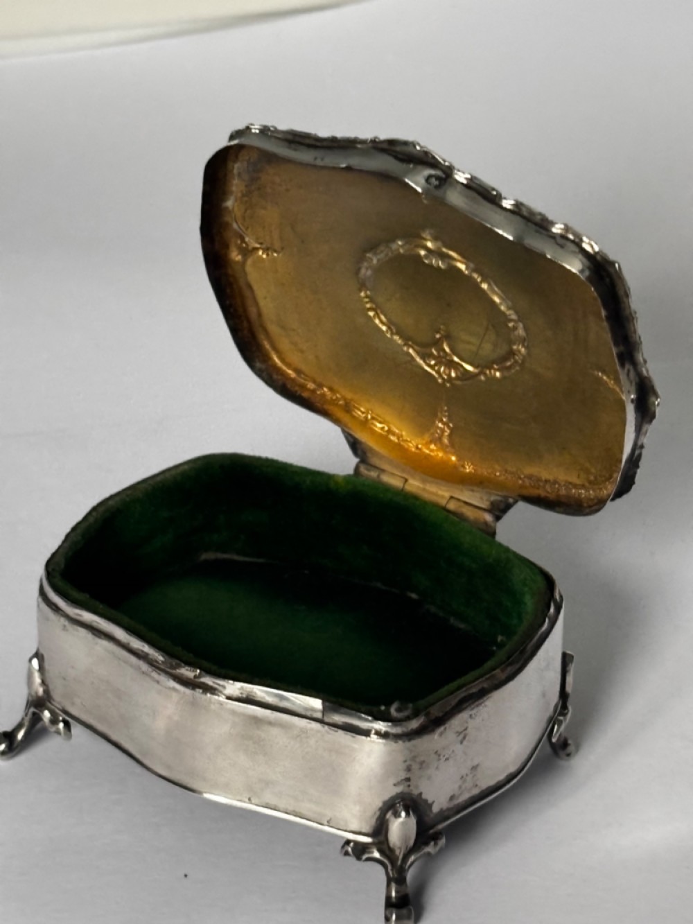 An open George V sterling silver trinket box featuring green velvet lining, gilt lid interior, and cabriole feet.