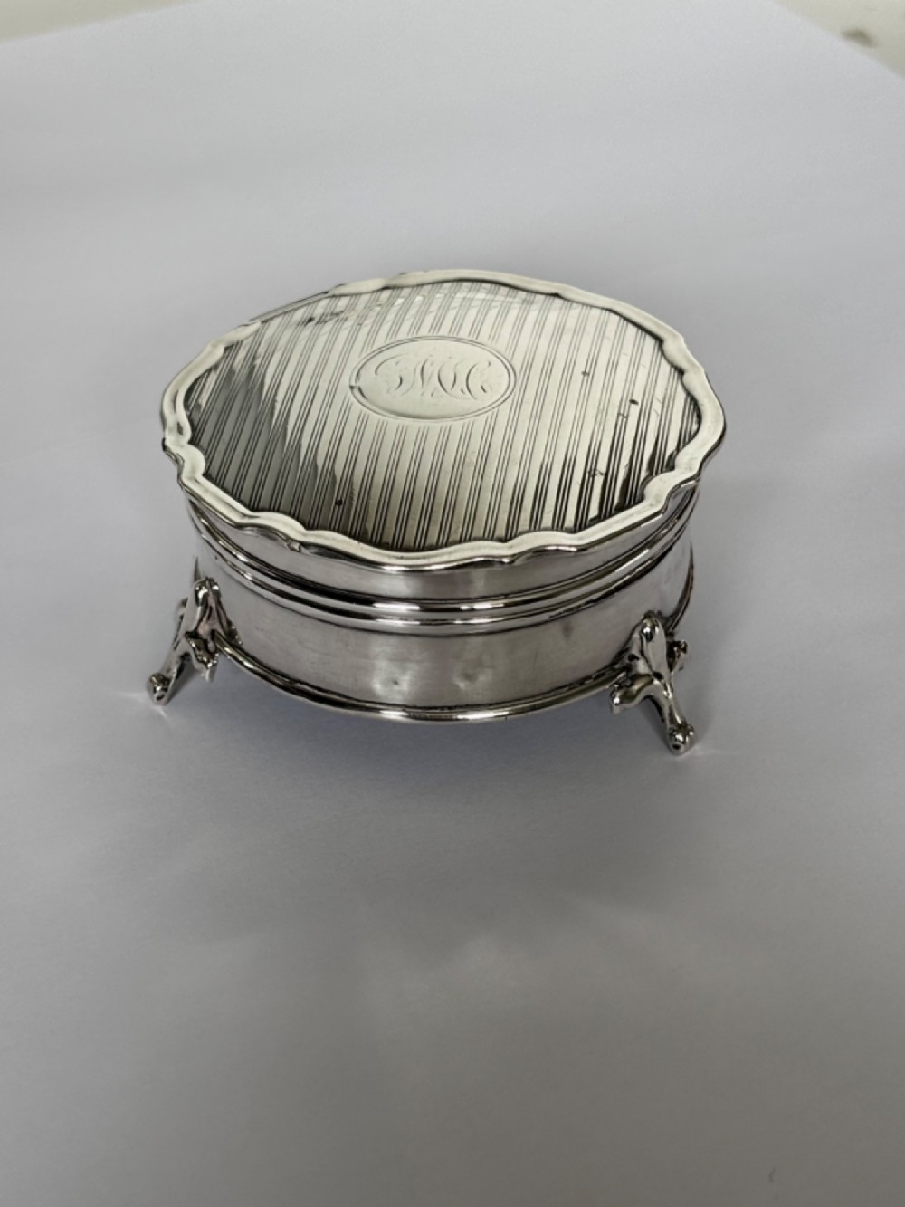 A circular George V sterling silver trinket box featuring engine-turned lid decoration, an engraved central cartouche, and four short pad feet.