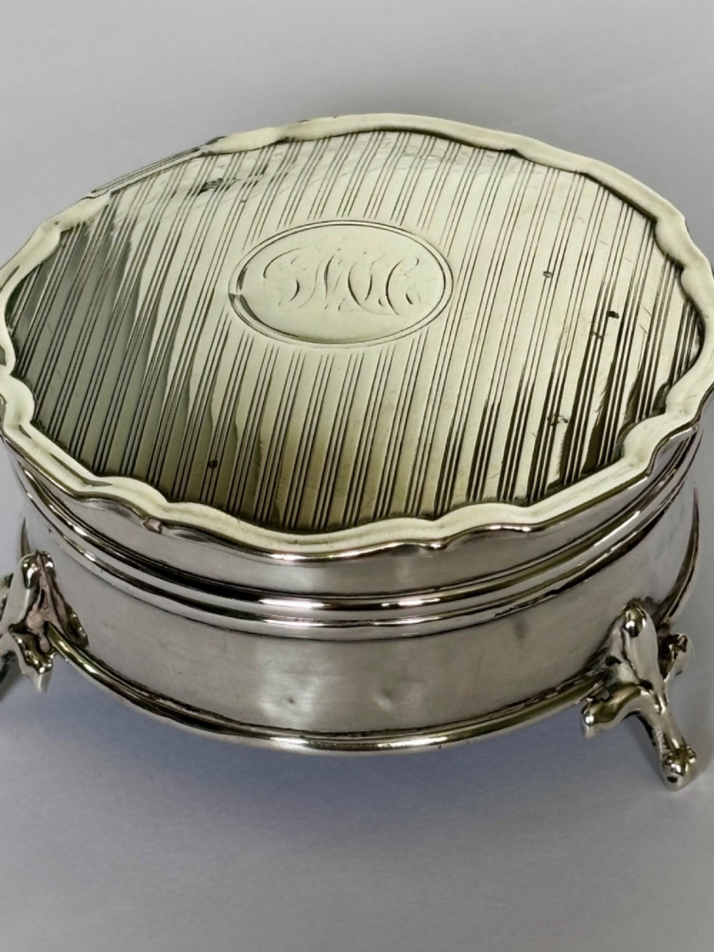 A circular George V sterling silver trinket box featuring engine-turned lid decoration, an engraved central cartouche, and four short pad feet. View II