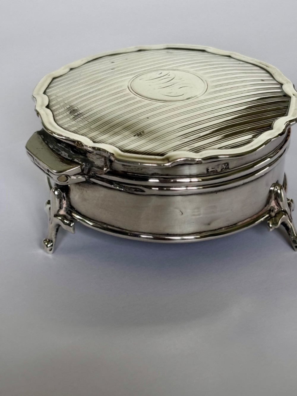A circular George V sterling silver trinket box featuring engine-turned lid decoration, an engraved central cartouche, and four short pad feet. View V