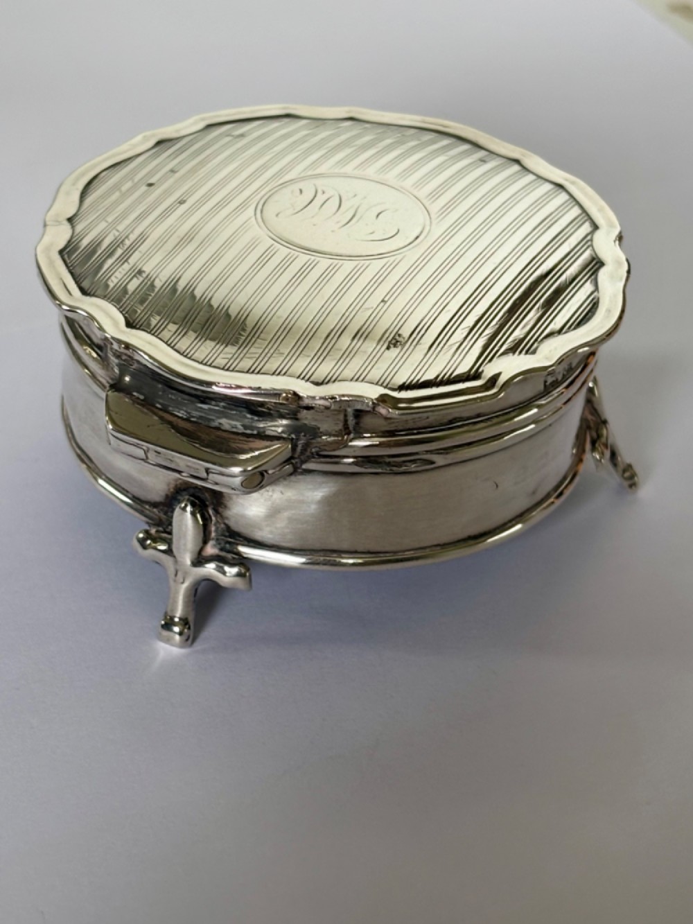 A circular George V sterling silver trinket box featuring engine-turned lid decoration, an engraved central cartouche, and four short pad feet. View VII